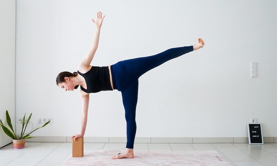 A person is performing a half-moon yoga pose with the assistance of a block in a bright, minimalistic room.