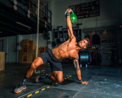 A shirtless, fit man with tattoos performs a kettlebell exercise in a well-equipped gym.